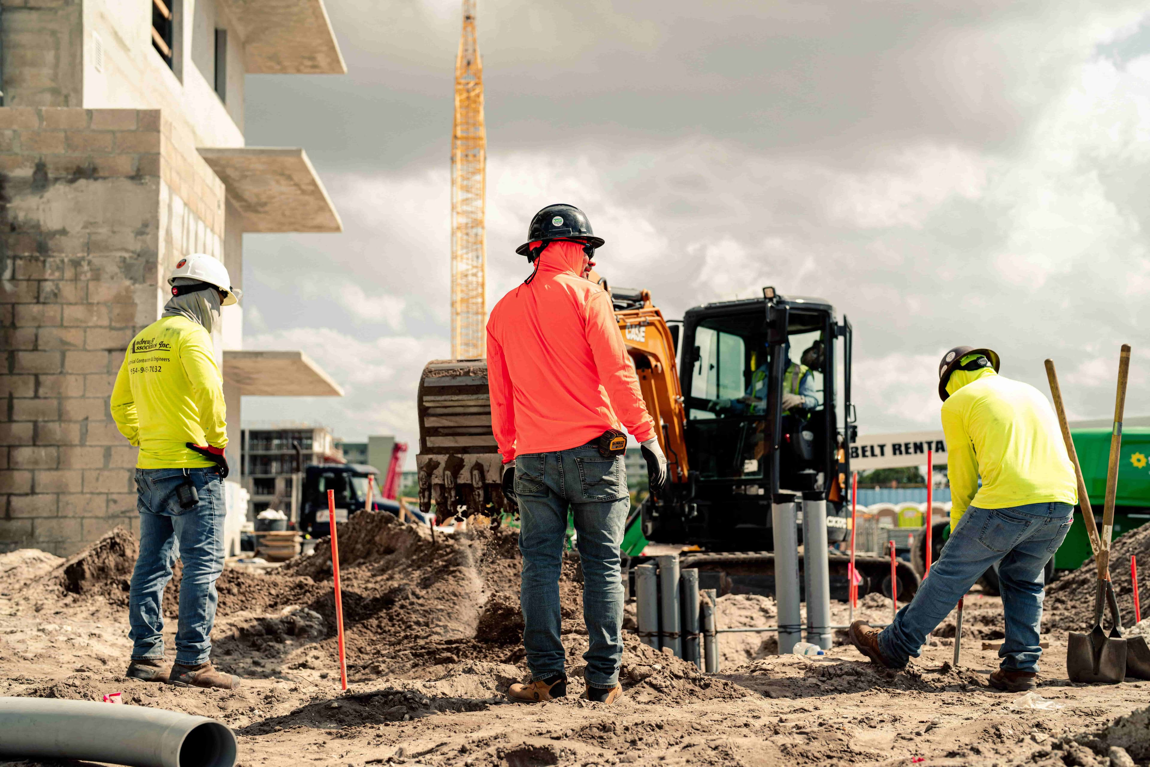 Construction crew working on a sandy jobsite with excavator and high visibility safety gear visible in the background.